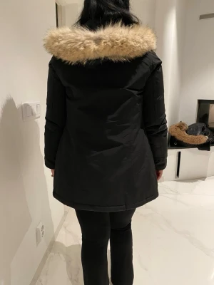 Svart parkas med fuskpäls - Snygg svart parkas med huva och beige fuskpäls runt kanten. Jackan har dragkedja och knappar framtill samt en rak passform som går ner över höfterna. Perfekt för kalla dagar och ger en stilren look. Materialet ser ut att vara vattenavvisande syntet.