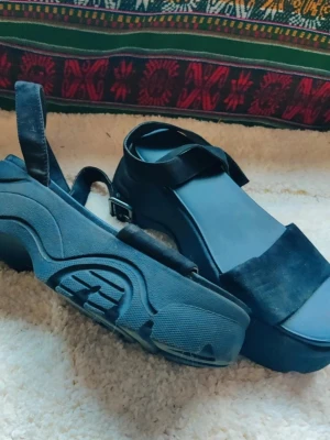 Svarta platåsandaler med remmar chunky sandals 38 - Chunky svarta sandaler med breda remmar och kraftig platåsula. Justerbart spänne vid ankeln och öppen tå. Tillverkade i mockaimitation och gummi för en cool och edgy look.