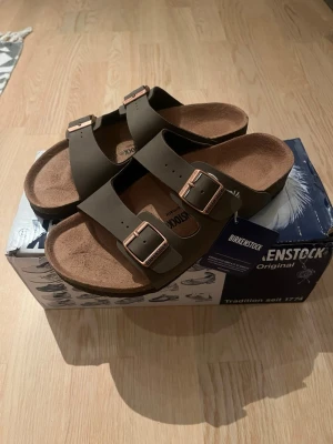 Bruna Birkenstock Arizona sandaler - Säljer ett par klassiska Birkenstock Arizona sandaler i brun färg med två justerbara remmar och metallspännen i kopparfärg. Fotbädden är i mocka och sulan är ergonomiskt utformad för extra komfort. Perfekta för en avslappnad och trendig stil. Dem är lite breda i storlek 
