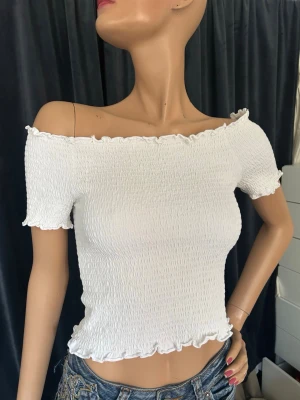 Vit offshoulder topp - Trendig vit oneshoulder topp med smockad struktur och korta ärmar. Toppen har volangkant vid halsringning, ärmslut och nederkant för en söt touch. Passar small!! Perfekt att styla med jeans eller kjol för en fräsch look.