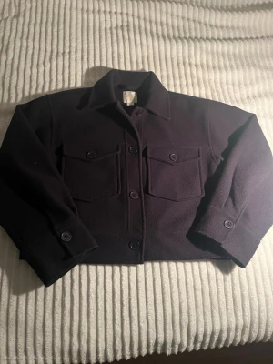 Marinblå kort kappa med fickor från Gina! - Stilren marinblå kappa med två  bröstfickor och knappar framtill. Kappan har klassisk krage och är tillverkad i ett tjockare, mjukt material som liknar ull. Perfekt för lager på lager och ger en cool, avslappnad look. Storlek 170 men passar absolut xs/s