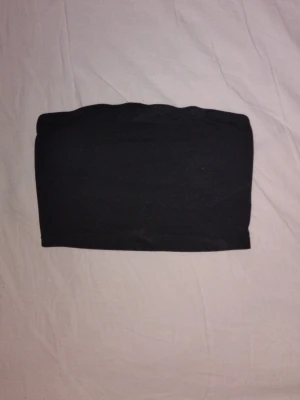 Svart bandeau tubtopp - En enkel och stilren svart bandeau tubtopp i stretchigt material. Toppen är axelbandslös och har en tight passform som sitter snyggt på kroppen. Perfekt att matcha med höga jeans eller kjol för en trendig look.