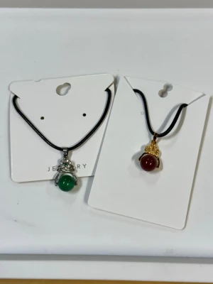 Två halsband med jade berlocker  - Säljer två halsband med svarta band och runda jade berlocker. Ena halsbandet har en grön hade med silverfärgad infattning, det andra har en vinröd jade med guldfärgad infattning och dekorativa detaljer. Perfekta accessoarer för att lyfta din stil.