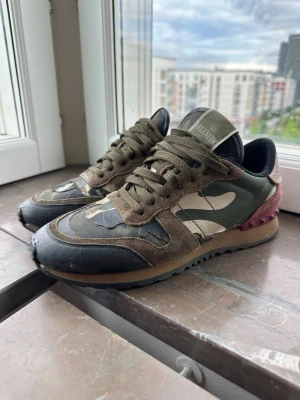 camo sneakers i grönt och brunt - Snygga sneakers med camomönster i grönt, brunt och beige. Skorna har detaljer i mocka och läder, samt en cool vinröd häl. Klassisk låg modell med snörning och robust gummisula. Perfekta för dig som vill sticka ut med stil.