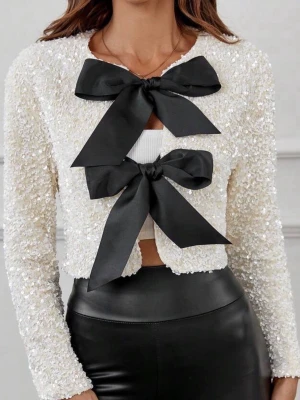 Paljettkofta med stora svarta rosetter - Glittrig paljettkofta i offwhite med två stora svarta rosetter framtill. Kort modell med lång ärm och öppen front. Perfekt för att lyfta en outfit med extra glamour och statement-detaljer.