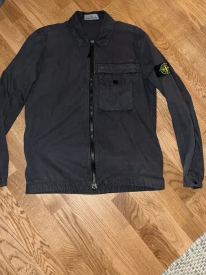 Svart overshirt från Stone Island - Snygg svart overshirt från Stone Island med dragkedja framtill, bröstficka med knapp och den klassiska logotypen på ärmen. Tillverkad i bomull med en avslappnad passform och skjortkrage. Perfekt lager på lager-plagg för en trendig streetwear-look.
