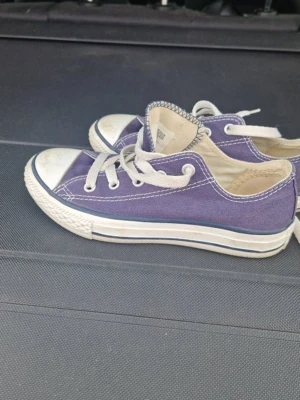 Lila Converse All Star låga sneakers - Klassiska Converse All Star sneakers i lila canvas med vita gummisulor och tåhätta. Skorna har vita snören och detaljer i vitt och blått. Perfekta för en avslappnad och cool stil. Ikonisk logga på innersulan och tålig design.