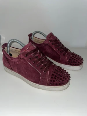 Christian Loubotin - Snygga vinröda sneakers i mocka fråna Christian Loubotin med ikoniska nitar på tån och vit sulor. Skorna har matchande vinröda skosnören och en låg silhuett. Perfekta för dig som vill sticka ut med en lyxig och edgy stil