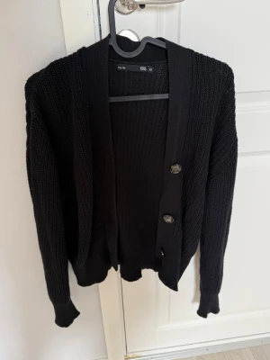 Svart stickad kofta med knappar - Säljer en svart stickad kofta från H&M med tre stora knappar framtill. Koftan har lång ärm, ribbade muddar och en avslappnad passform. Perfekt att slänga över en t-shirt eller linne för en enkel och snygg look.