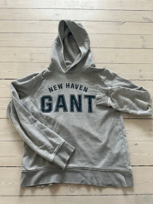 Grå hoodie från GANT med tryck - Snygg grå hoodie från GANT med stor huva och trycket 'NEW HAVEN GANT' i mörkblått framtill. Klassisk modell med ribbade muddar vid ärmslut och nederkant. Tillverkad i mjukt sweatshirtmaterial, perfekt för en avslappnad stil.