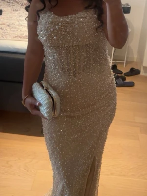 Champagnefärgad långklänning med pärlor - Elegant långklänning i champagnefärg täckt av pärlor och paljetter. Klänningen har en figurnära passform, slits framtill och är ärmlös med axelbandslös design. Perfekt för bal eller festliga tillfällen. Klänningen har varit skräddad även och den var köpt för runt 2000, etiketten på finns fortfarande kvar på klänningen. Den har en inbyggd bh och en inbyggd korsett inuti. 