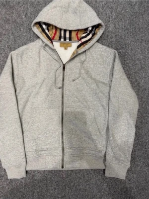 Grå zip hoodie från Burberry - Snygg grå zip hoodie från Burberry med klassiskt rutigt foder i huvan. Hoodien har dragsko i huvan, ribbade muddar och mjukt material på insidan. Perfekt för en avslappnad och trendig look.