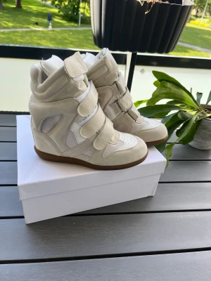 Beiga wedge sneakers Isabel marant - Snygga beiga sneakers med inbyggd kilklack och flera breda kardborreband. Skorna har en mix av mocka och textil, samt vit detalj på tån och sidorna. Perfekta för dig som vill ha lite extra höjd och en cool streetstil.