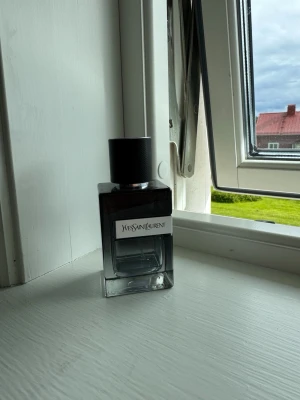 Yves Saint Laurent parfym herr - Snygg parfymflaska från Yves Saint Laurent med stilren, fyrkantig design. Flaskan är i glas med mörkgrå till svart toning och har en svart, räfflad kork. Märkets logga syns tydligt på framsidan. Perfekt för dig som gillar exklusiva dofter.