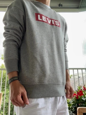 Grå sweatshirt från Levi's - Säljer en klassisk grå sweatshirt från Levi's med det ikoniska röda logotrycket på bröstet. Tröjan har rund halsringning, ribbade muddar vid ärmslut och nederkant samt en avslappnad passform. Perfekt till jeans eller shorts för en clean och enkel stil. Nästan aldrig använd