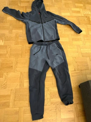 Nike tech tracksuit blå  - Nike Tech Fleece tracksuit i två nyanser av grått med svarta detaljer. Byxorna har dragsko i midjan, muddar vid bensluten och en liten Nike-logga på låret. Tillverkade i mjukt och stretchigt material som är perfekt för chill eller träning.