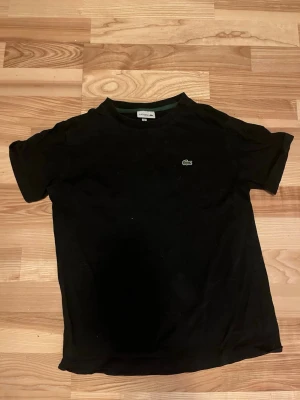 Svart t-shirt från Lacoste - Klassisk svart t-shirt från Lacoste med den ikoniska gröna krokodilloggan broderad på bröstet. T-shirten har rund hals och korta ärmar, perfekt för en clean och enkel stil. Tillverkad i mjuk bomull för skön komfort.