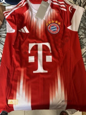 Bayern München matchtröja Adidas röd - Snygg röd matchtröja från Adidas för FC Bayern München med klubbmärke och fem stjärnor på bröstet. Stora vita detaljer och logga framtill, samt FC Bayern München-tryck på ryggen. Tillverkad i lätt och ventilerande material, perfekt för fotboll.