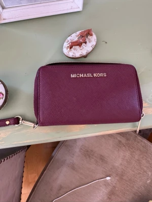 Vinröd plånbok från Michael Kors - Snygg vinröd plånbok från Michael Kors i tåligt saffianoläder. Plånboken har dragkedja runtom, flera kortfack, myntfack med dragkedja och ett fönster för ID eller foto. Guldfärgade detaljer och avtagbar handledsrem gör den både praktisk och stilren.