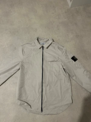 Beige overshirt från Stone Island - Snygg beige overshirt från Stone Island med svart dragkedja framtill och klassisk patch på ärmen. Overshirten har en bröstficka med lock och är tillverkad i ett lätt material som passar perfekt för lager-på-lager. Enkel, stilren design med modern touch.
