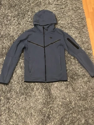 Blå Nike Tech Fleece hoodie - Snygg blå Nike Tech Fleece hoodie med dragkedja framtill och huva. Har svarta detaljer över bröstet och en ficka med dragkedja på ena ärmen. Tillverkad i mjukt och stretchigt material som ger en modern och sportig look. Perfekt för dig som gillar streetwear. Gamla modellen av Nike tecks som är lite snyggare