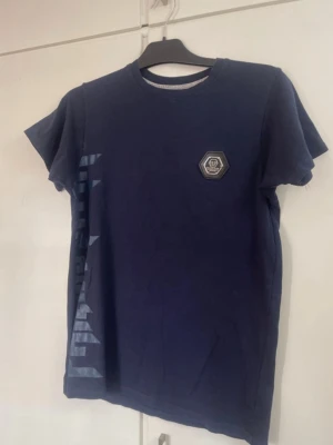 Blå Philipp Plein t-shirt med dödskalle - Snygg mörkblå t-shirt från Philipp Plein med stort dödskalletryck och texten 'PLEIN' på ryggen. Framtill finns en liten logga på bröstet och tryck längs sidan. Klassisk rund hals och korta ärmar. Perfekt för dig som gillar statement-plagg.