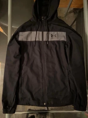 Svart vindjacka från Under Armour - Snygg svart vindjacka från Under Armour med grått bröstparti och logga. Jackan har huva med dragsko, hel dragkedja framtill och två sidofickor. Tillverkad i lätt och tåligt material med meshfoder för extra komfort.