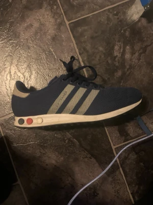 Marinblå Adidas sneakers med retrolook - Snygga marinblå sneakers från Adidas med klassiska tre vita ränder på sidan. Skon har en retrovibb med färgade detaljer på sulan och ovandel i mesh-tyg. Låg modell med vita och röda knappar på sidan av sulan. Perfekt för dig som gillar vintage-stil.