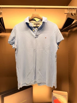 Tommy Hilfiger - Snygg ljusblå pikétröja från Tommy Hilfiger med klassisk krage och knappar framtill. Liten broderad logga på bröstet och custom fit-passform. Perfekt till jeans eller shorts för en clean och stilren look.
