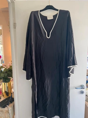 Svart långklänning från By Malina - Stilren svart långklänning från By Malina med vita kontrastkanter längs v-ringningen, ärmsluten och nederkanten. Klänningen har lång ärm och en avslappnad passform, perfekt för en chic och enkel look. Oanvänd 