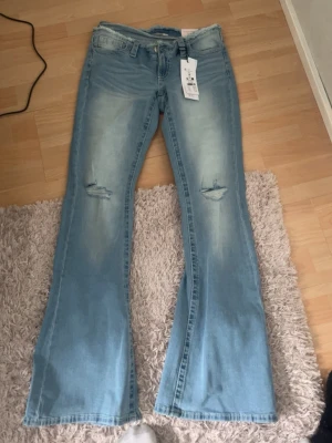 Ljusblå bootcut jeans med no waist - Säljer ett par ljusblå bootcut jeans med slitningar på knäna och snygg tvätt. Jeansen har klassiska fem fickor, låg midja och utsvängda ben. Perfekta för dig som gillar retrostil och vill ha ett par jeans som sticker ut.