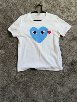 Vit t-shirt Comme des Garçons Play - Vit t-shirt från Comme des Garçons Play med stort blått hjärta och ett litet rött hjärta med ögon på bröstet. Klassisk rund hals och korta ärmar. Perfekt för dig som gillar streetwear och vill sticka ut med en ikonisk design.