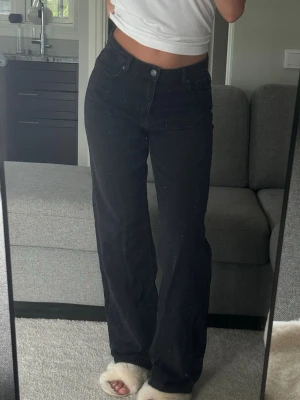 Svarta raka jeans med glitterdetaljer - Säljer ett par svarta jeans med raka ben och diskreta glitterdetaljer. Jeansen har hög midja och klassisk femficksmodell. Perfekta för dig som vill ha en trendig och bekväm look med lite extra bling.
