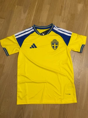 Sveriges vm tröja M - Gul svensk landslagströja från Adidas med blå detaljer och vita ränder på axlarna. Broderad landslagssköld på bröstet och 'Sverige' tryckt i nacken. Tillverkad i lätt och ventilerande material, perfekt för fotboll eller träning.