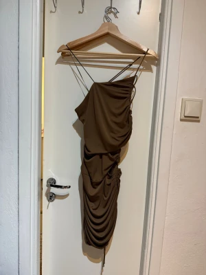 Brun asymmetrisk omlottklänning - Säljer en brun kortklänning med asymmetrisk design och tunna axelband. Klänningen har omlottdetaljer och rynkade partier längs sidan för en snygg draperad effekt. Perfekt för dig som gillar trendiga och unika plagg.