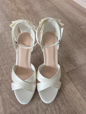 Sandalett i satin med rosett bak och kryssrem 37 - Sandalett i färgen Ivory med öppen tå, smala remmar som korsas framtill och en elegant rosett bak på hälen. Skon har en smal klack och justerbar ankelrem med spänne. Perfekta för dig som vill ha en chic och feminin look.