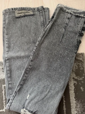 Grå jeans med slitningar och stänk - Snygga grå jeans med rå kant i midjan, slitningar vid benslut och ficka samt coola vita färgstänk över hela tyget. Knappgylf med svarta knappar ger en extra detalj. Perfekta för dig som gillar en trendig och avslappnad stil.