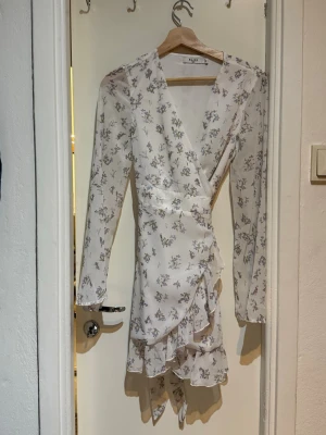 Vit blommig omlottklänning NA-KD - Söt vit omlottklänning från NA-KD med blommigt mönster i lila och grönt. Klänningen har långa ärmar, volangdetaljer och v-ringad hals. Perfekt för sommarens alla tillfällen!