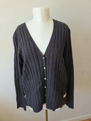 Odd Molly Mörkgrå kabelstickad kofta L (3) - Mysig mörkgrå kabelstickad kofta med v-ringning och vita knappar framtill från Odd Molly. Koftan har långa ärmar och ribbade muddar vid ärmslut och nederkant. Perfekt att slänga över en t-shirt för en avslappnad look.
