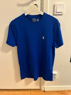 Blå t-shirt från Polo Ralph Lauren - Klassisk blå t-shirt från Polo Ralph Lauren med gul broderad logga på bröstet. Modellen är kortärmad och har rund halsringning. Tillverkad i mjuk bomull för en bekväm känsla och har en normal passform.