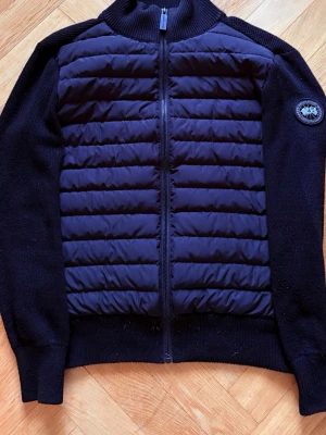 Svart cardiganjacka från Canada Goose - Snygg svart cardiganjacka från Canada Goose med quiltad front och stickade ärmar samt rygg. Jackan har dragkedja framtill och en rund halsringning. Märkets logga sitter på vänster ärm. Perfekt för lager-på-lager och streetstyle.