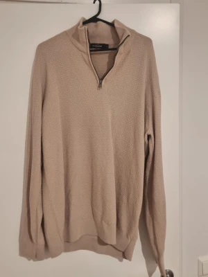 Beige stickad halvzip från Dressman - Snygg beige stickad tröja från Dressman med halv dragkedja och hög krage. Tröjan har diskret strukturmönster, ribbade muddar och är perfekt för lager på lager. Materialet känns mjukt och bekvämt.