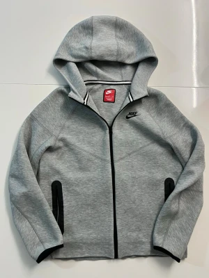 Grå Nike tech kofta från Nike - Snygg grå kofta från Nike med dragkedja framtill och svart Nike-logga på bröstet. Koftan har två svarta fickor med dragkedja och en stor huva. Tillverkad i mjukt sweatshirtmaterial, perfekt för en sportig och avslappnad stil.