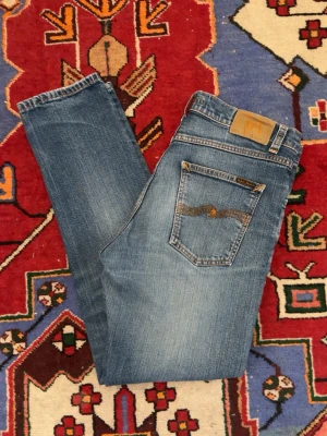 Blå jeans från Nudie Jeans - Säljer ett par klassiska blå jeans från Nudie Jeans med snygg tvätt och diskreta slitningar. Modellen har raka ben och fem fickor, samt Nudie Jeans ikoniska broderi på bakfickan. Perfekta för en avslappnad och trendig stil.