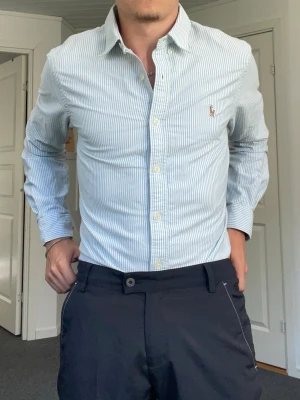 Skjorta från Polo Ralph Lauren - Säljer en ljusblå och vit randig skjorta från Polo Ralph Lauren i slim fit. Skjortan har klassisk krage, knappar framtill och den ikoniska broderade loggan på bröstet. Tillverkad i bomull och har långa ärmar.