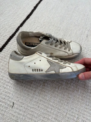Vita Golden Goose Superstar sneakers - Golden Goose Superstar sneakers i vitt läder med grå stjärndetalj på sidan, grå mockadetaljer vid häl och grå skosnören. Klassisk låg modell med slitna detaljer för en cool och avslappnad look. Perfekta för dig som gillar streetstyle och unika skor.