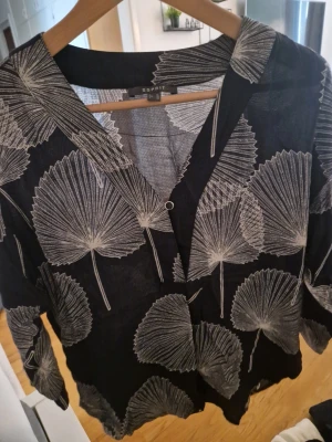 Svart blus med vita blad från Esprit - Snygg svart blus från Esprit med vita grafiska bladmönster. Blusen har v-ringning, knapp framtill och är tillverkad i ett tunt, lätt material som känns luftigt. Perfekt för dig som gillar naturinspirerade prints och vill ha en avslappnad men trendig look.