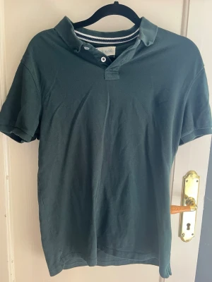 Mörkgrön piké från Pier One - Mörkgrön pikétröja från Pier One med klassisk krage och två knappar vid halsen. T-shirten är kortärmad och har en enkel, stilren design som passar till det mesta. Tillverkad i bomull för en skön känsla.