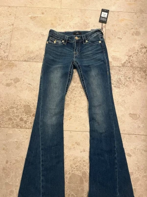 True Religion Joey Low Rise Flare Jeans - Blå jeans från True Religion i modellen Joey Low Rise Flare. Jeansen har låg midja, utsvängda ben och fickor med lock. Tillverkade i klassisk denim med synliga kontrastsömmar och cool vintagekänsla. Perfekt för dig som gillar en trendig och avslappnad look. Oanvända. Lappen kvar. Nypris 1299kr