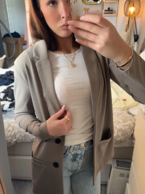 Beige oversized kavaj med fickor - Snygg beige oversized kavaj med två framfickor och svarta knappar. Kavajen har klassisk krage och är tillverkad i ett mjukt, tunt material som ger en avslappnad look. Perfekt att slänga över en t-shirt och jeans för en trendig stil.